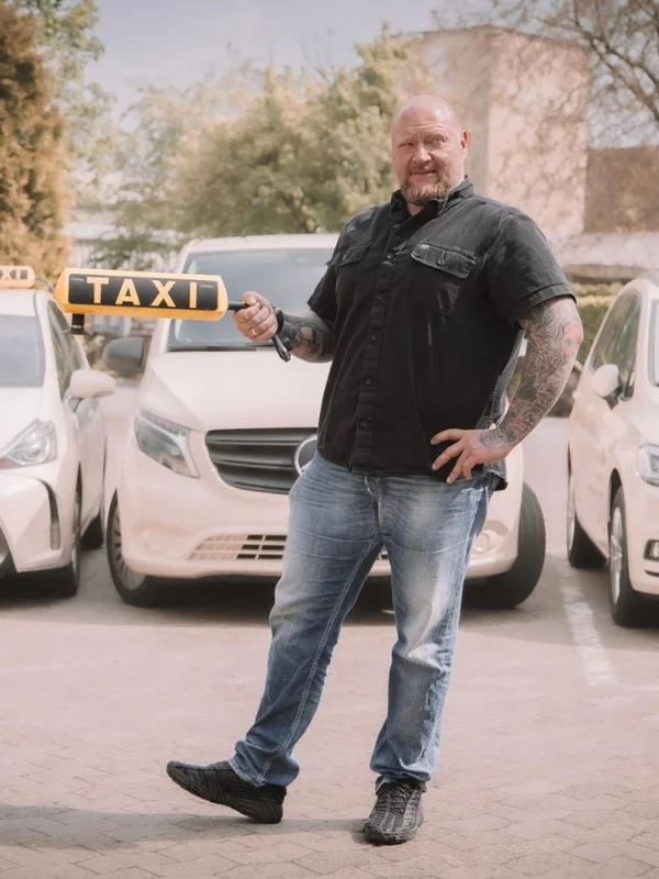 Kräftiger Mann steht vor mehreren Taxis und hält ein Taxi-Schild, repräsentiert eine organisierte Taxengemeinschaft.